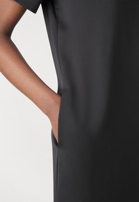 Rochie neagră dintr-un material neted, cu o croială lejeră, dotată cu un buzunar lateral și mâneci scurte. Textura pare moale și elegantă.
