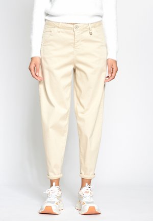 Person trägt beigefarbene hochgekrempelte Hose, weißen strukturierten Pullover und weiße Sneakers mit orangefarbenen Akzenten, steht vor weißem Hintergrund.