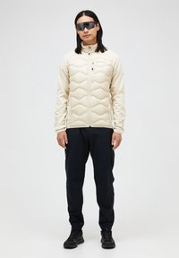 Lys beige polstret jakke med et kviltet mønster, lynlåslukning og høj krave, parret med sorte bukser og sorte sneakers.