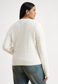 Kobieta o długich ciemnych włosach nosi biały sweter w warkocze i niebieskie dżinsy, stoi tyłem, a jej głowa jest widoczna z profilu.