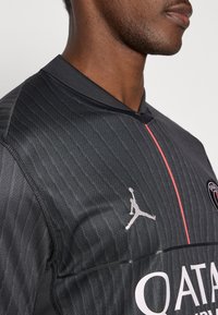 Fekete sportmez texturált csíkokkal, rövid gallérral és rózsaszín részletekkel. Szürke Jumpman logóval és "QATAR" felirattal ellátva.