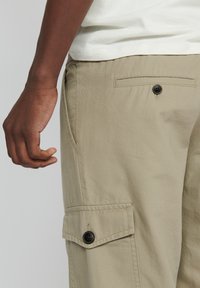 Beige Cargo-Hosen aus einem Baumwollmischgewebe, mit Seitentaschen, einer mit Knopf verschlossenen Tasche und geradem Schnitt. Sichtbare Struktur auf der Oberfläche.