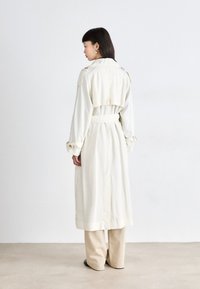 Manteau tranché crème avec une taille ceinturée, des poignets boutonnés et une fente au dos ; porté sur un pantalon beige clair ; texture lisse, design ajusté.