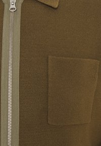 Pull marron à fermeture éclair avec une texture côtelée, doté d'une fermeture éclair verte contrastante et d'une poche avant sur le côté gauche.