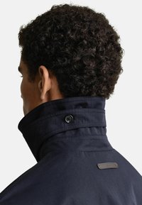 Granatowa kurtka z kołnierzem stojącym, z dwoma czarnymi guzikami. Materiał wydaje się gładki z delikatnym połyskiem i detalem logo na kołnierzu.