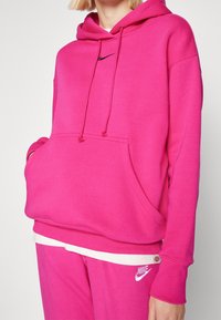Felpa con cappuccio rosa, dotata di tasca frontale, coulisse e logo Nike nero. Tessuto morbido e polsini a coste, abbinata a pantaloni rosa coordinati.