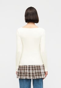 Γυναίκα με κοντά σκούρα μαλλιά, που φοράει λευκό τοπ με μακριά μανίκια πάνω από καρό καφέ φούστα και μπλε τζιν, με την πλάτη γυρισμένη σε απλό φόντο.