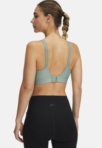 Športni nedrček v svetlo zeleni tkanini z širokimi naramnicami in zaponko na hrbtu. V kombinaciji z visokimi črnimi legicami.