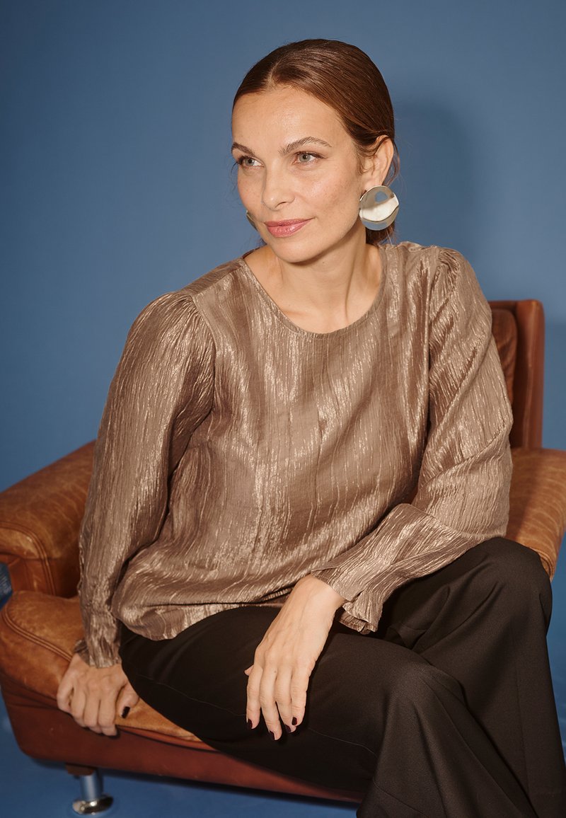 Pronksikarvaline pluus pika varrukaga, ümar kaelus; kombineeritud mustade pükstega. Model istub pruuni nahast diivani peal sinise tausta ees.