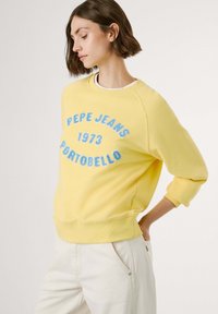 Geltonas džemperis su apvalia apykakle, puošnintas mėlyna siuvinėta išraiška "PEPE JEANS 1973 PORTOBELLO." Įprastas siluetas ir pūkuoti rankogaliai.