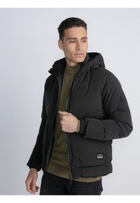 Chaqueta de plumas negra con capucha, que presenta un cierre frontal, puños elásticos y un pequeño parche de la marca en el lado inferior izquierdo. Combinada con una parte superior verde oliva.