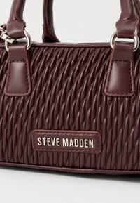 Mala de couro bordô com pregas diagonais texturizadas, alças duplas, anéis prateados e placa de logo em metal "Steve Madden" na frente.