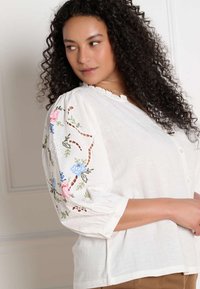 Vrouw met krullend haar, gekleed in een witte blouse met kleurrijke bloemenborduursels op opgeblazen mouwen, kijkt over haar schouder.