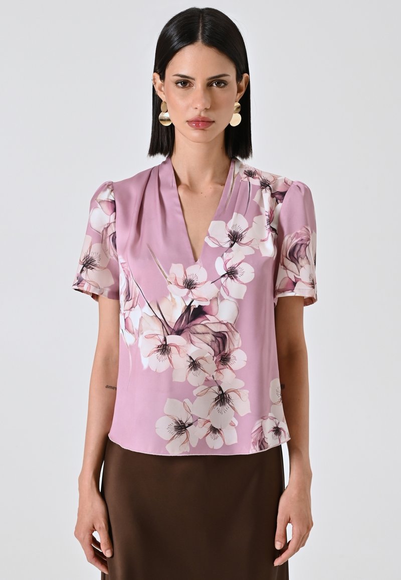 Blusa floreale rosa in satin con maniche a sbuffo corte, caratterizzata da un profondo scollo a V e grandi fiori bianchi con accenti neri.
