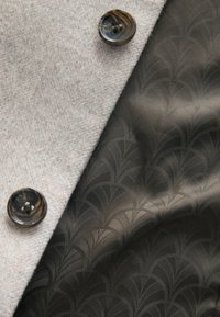 Lys grå filtstof med mørk mønstret foring, der har en rund, mørk knap med fire huller til fastgørelse.