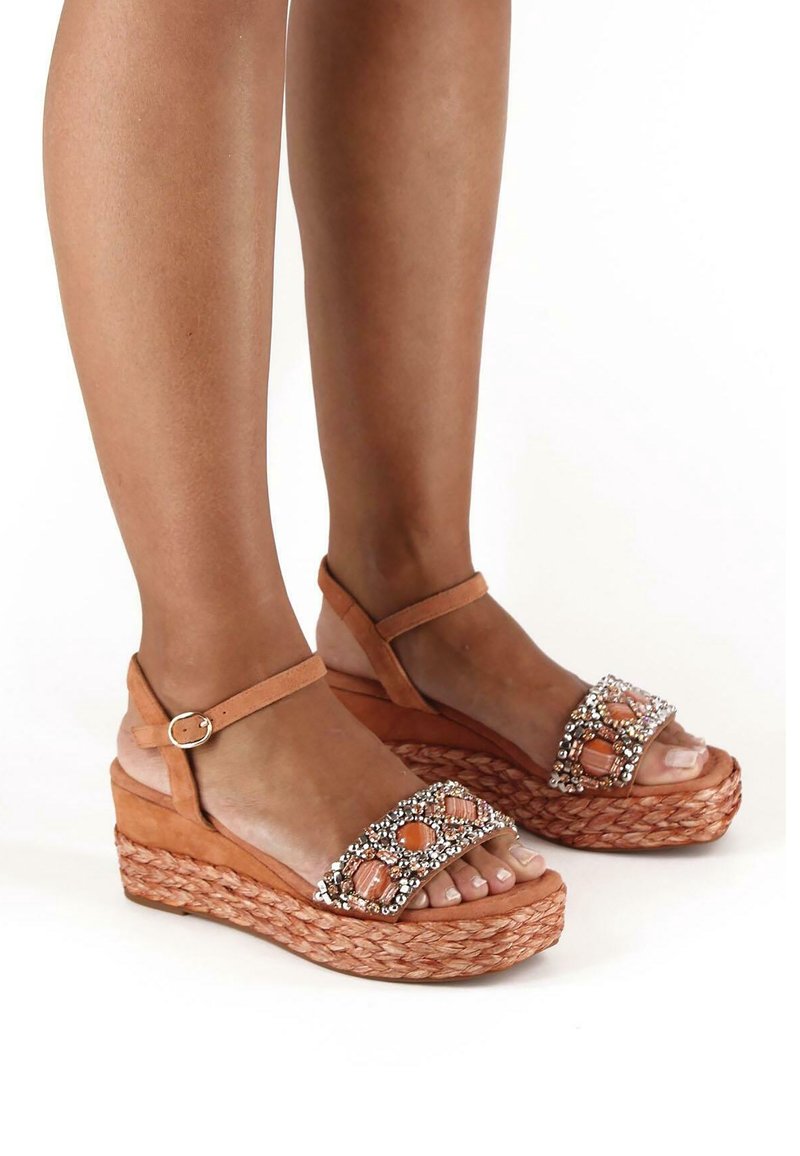 Pieds portant des sandales compensées couleur tan avec des semelles tressées et des brides ornées de bijoux, maintenues par des brides de cheville avec de petites boucles.