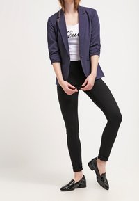 Granatowa marynarka na białym topie na ramiączkach z cekinowym napisem, zestawiona z czarnymi legginsami i błyszczącymi, czarnymi balerinkami. Gładki materiał, dopasowany krój.