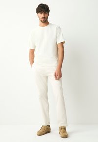 Top color beige chiaro a maniche corte, tessuto testurizzato; abbinato a pantaloni bianchi aderenti e scarpe in camoscio marrone. Design semplice e moderno.