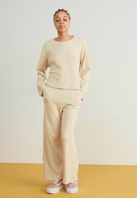Beige langermet topp og vide bukser laget av mykt stoff. Modellen har på seg lyserosa sko med hvite såler. Enkelt, avslappet design.