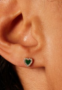 Goldene herzförmige Ohrringe mit einem grünen Edelstein in der Mitte, umgeben von kleinen klaren Steinen, vor einem Nahaufnahme eines hautfarbigen Ohrs.