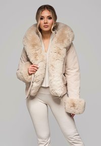 Beżowa pikowana kurtka z miękkim futrzanym kołnierzem i mankietami z futra ekologicznego, zapinana na zamek błyskawiczny, o gładkiej fakturze. Noszona na dopasowanym stroju.