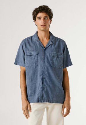 Giovane uomo con capelli ricci che indossa una camicia blu a maniche corte con bottoni e tasche sul petto, e pantaloni color crema, in piedi davanti a uno sfondo semplice.