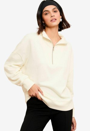 Cremefarbene zip-up Sweatjacke mit Stehkragen, langen Ärmeln und gerippten Bündchen. Kombiniert mit schwarzen, figurbetonten Hosen.
