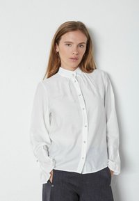 Hvid skjorte med høj krave, lange ærmer, let glans, tekstureret stof og guldtonede knapper ned langs fronten og ved manchetterne.