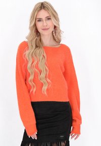 Pull en tricot orange vif avec une texture côtelée et des manches larges, associé à une jupe en daim noire avec des accents de franges.