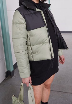 Personne portant une robe noire et une doudoune bicolore gris-vert, tenant un sac assorti gris-vert par sa lanière.