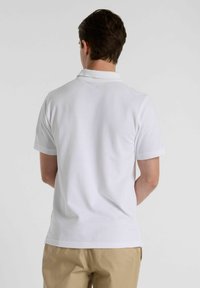 Camisa polo branca de mangas curtas com colarinho. Textura suave e corte reto, com um design clássico e acabamento simples. Sem logótipos visíveis.