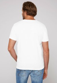 White cotton t-shirt with short sleeves and a crew neck, viewed from the back, paired with blue denim jeans. Simple, plain design.