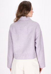 Lilla cropped jakke med en tekstureret stof, høj krave og lange ærmer. Har et subtilt vævet mønster og en lige kant.