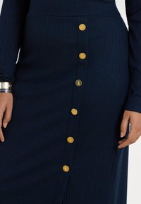 Saia azul-marinho com textura canelada, apresentando botões dourados destacados na frente, com um corte ajustado e uma pequena abertura lateral. Textura suave em toda a peça.