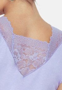 Fioletowa bluzka z dekoltem w kształcie litery V. Dekolt ozdobiony jest misternymi koronkowymi detalami z kwiatowymi wzorami i falbaniastymi brzegami.