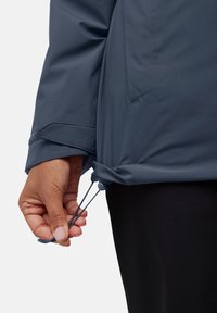 Blauwe jas van een glad, lichtgewicht materiaal met een verstelbare koord aan de onderrand. Close-up van een hand die het koord aantrekt.