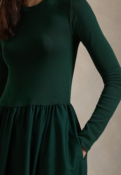 Grön klänning med långärmad i mjukt tyg, med en figursydd överdel och en veckad kjol. Inkluderar fickor och en rund halsringning.