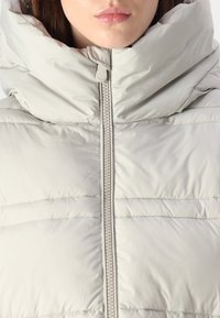 Lichtgrijze gewatteerde jas met een hoge, oversized kraag, voorzien van horizontaal gestikte secties en een centrale rits met een trekker.