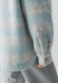 Veste chemise texturée bleu clair et crème avec une finition duveteuse, dotée d'un poignet à bouton et d'une ligne d'ourlet arrondie. Tissu doux et confortable.