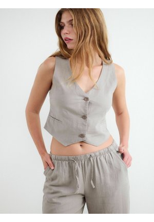 Kobieta z długimi włosami, ubrana w szarą kamizelkę bez rękawów zapinaną na guziki oraz pasujące spodnie z elastycznym pasem, ręce w kieszeniach, patrząca na bok.