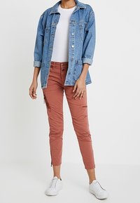 Person som bär en ljusblå jeansjacka över en vit skjorta, rostfärgade slim-fit byxor med dragkedjor och vita sneakers som står mot en vit bakgrund.