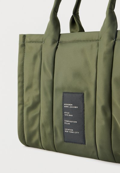 Τσάντα ώμου από νάιλον σε ελαιόχρωμο με επενδεδυμένα κάθετα πάνελ, δύο λαβές και μαύρη ετικέτα που αναγράφει τον σχεδιαστή Marc Jacobs, το στυλ, το υλικό και την τοποθεσία.