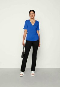 Top azul com mangas curtas, combinado com calças pretas ajustadas. Modelo usa saltos blocos brancos e segura uma pequena mala preta.