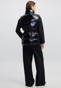 Chaleco acolchado negro con un acabado brillante, que presenta un cuello alto, cintura elástica y está acompañado de pantalones de pierna ancha negros. Textura suave.