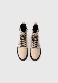 Bottines en cuir beige avec des lacets noirs et des semelles épaisses noires, présentées de dessus sur un fond blanc.