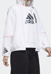 Hvid zip-up jakke med høj krave, elastisk kant og sort Adidas-logo. Har røde detaljer på skuldrene og lynlåsen. Båret over en hvid top.