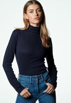 Haut col roulé bleu marine en tissu côtelé, à manches longues, associé à un jean bleu taille haute avec une fermeture à boutons et une coupe ajustée.