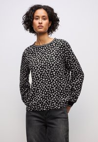 Donna con capelli ricci, indossa un top a maniche lunghe bianco e nero con motivo e jeans scuri, in piedi davanti a uno sfondo chiaro uniforme.
