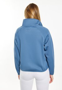 Blå fleecepola med hög krage, raglanärmar och en texturerad yta; har en subtil etikett vid bakre halslinje.