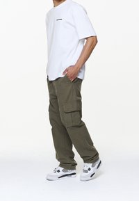Bílý bavlněný tričko s černým logem, ve spojení s olivově zelenými cargo kalhotami s několika kapsami a bílými teniskami s černými akcenty.